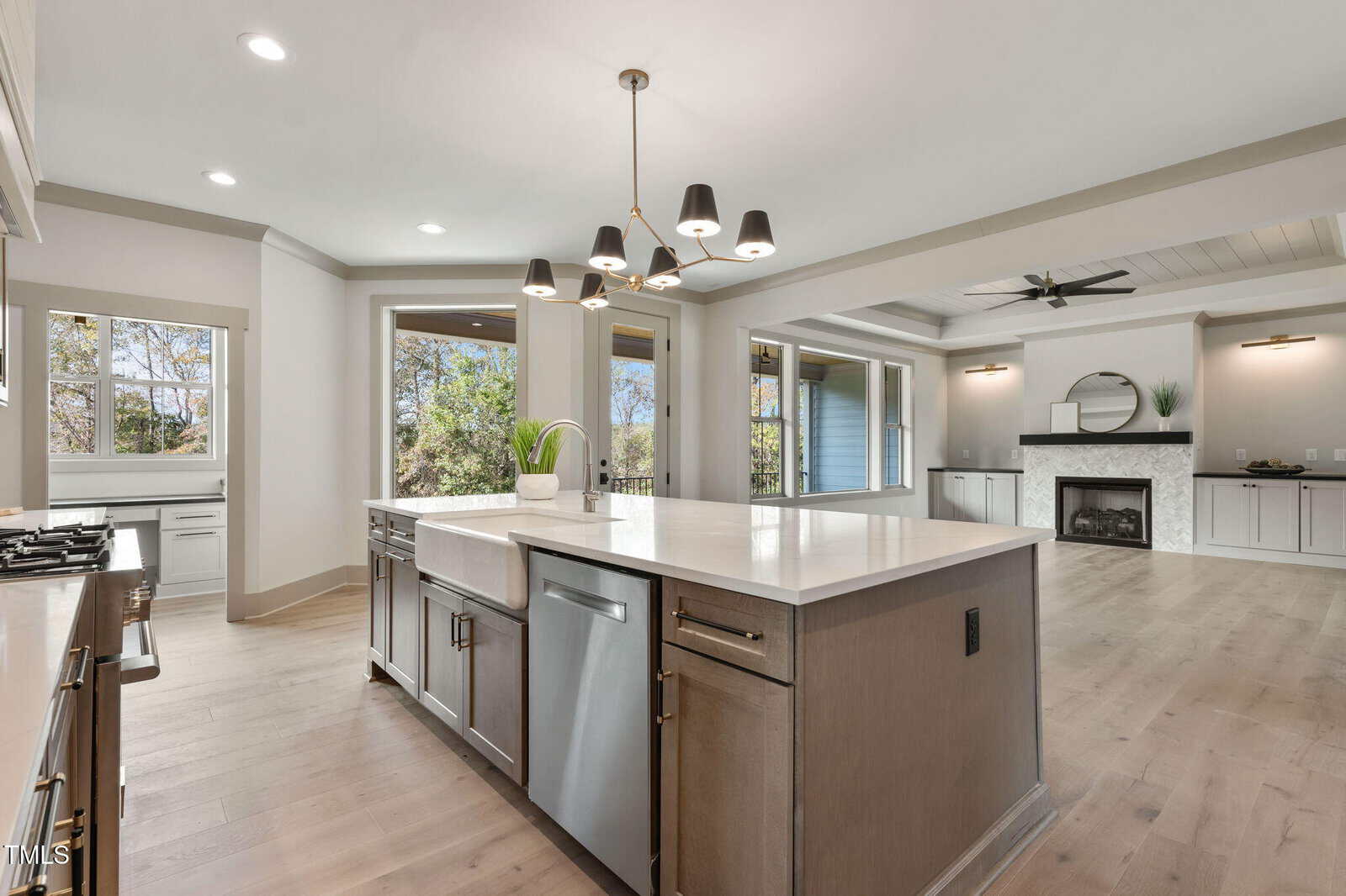 Modern kitchen with a large island, stainless steel appliances, pendant lighting, and open layout leading to a living room with a fireplace and large windows. Light wood floors and neutral colors throughout.
