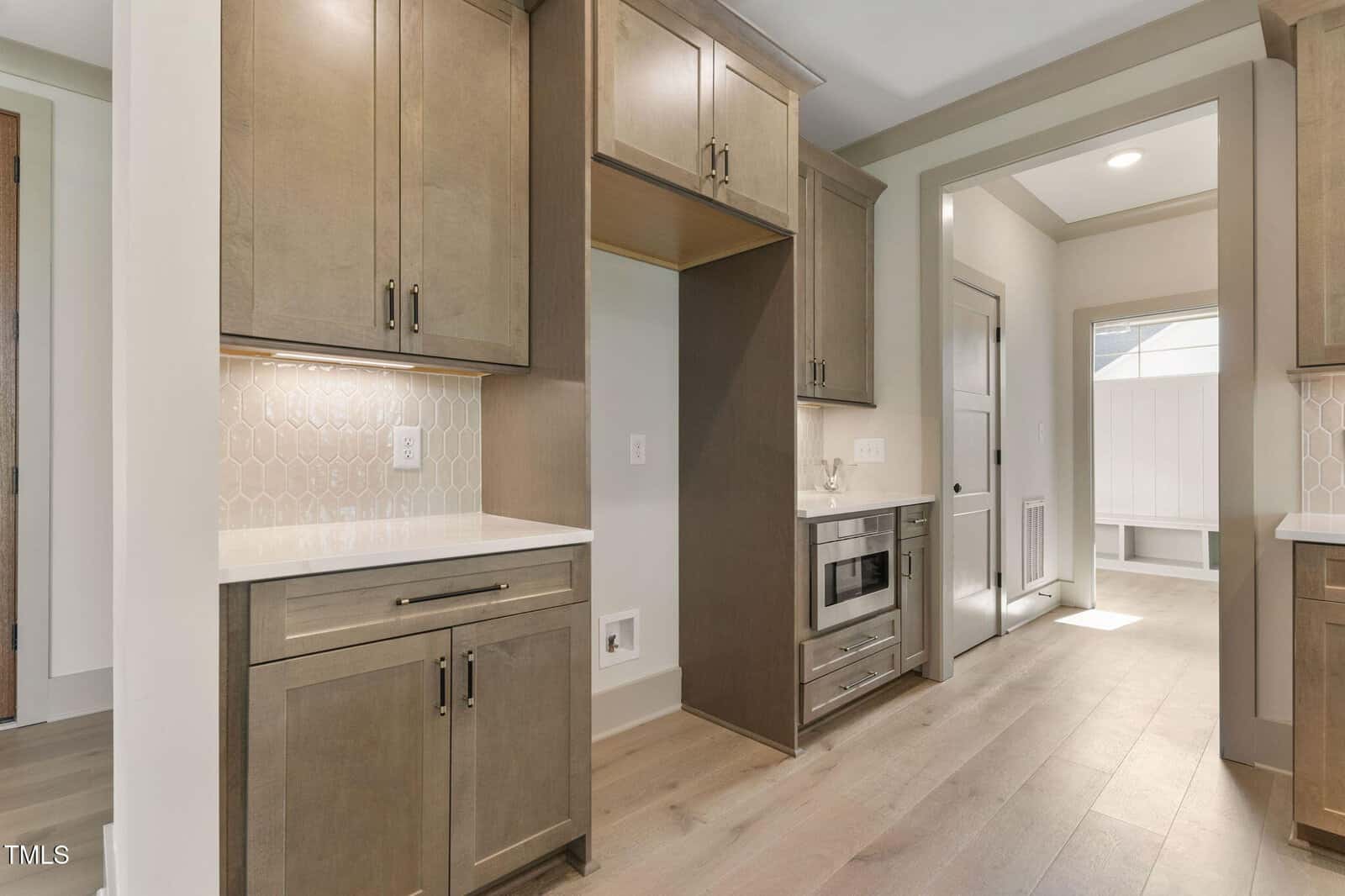Modern kitchen with wood cabinets, light countertops, a microwave, and oven built into lower cabinets. Light wood floors and a hallway leading to a mudroom with natural light visible in the background.
