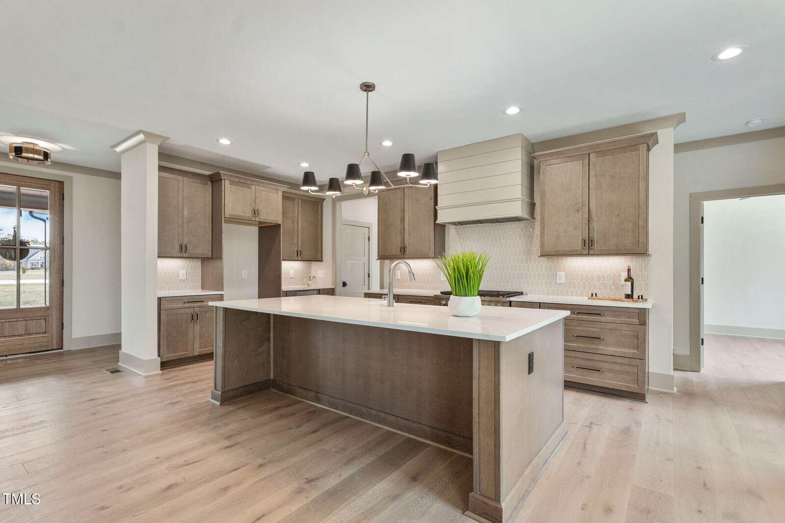 A modern kitchen with light wood cabinets, a large center island with a white countertop, a built-in oven, a range hood, and a contemporary black chandelier above the island. Light wood floors throughout.