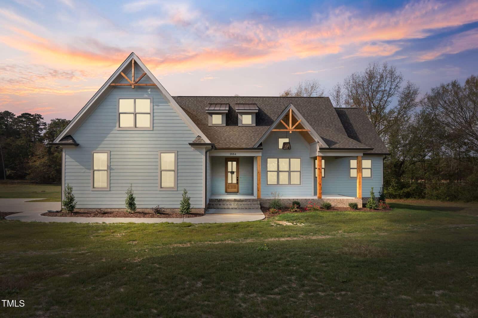 A modern two-story house with light blue siding, a dark gray roof, and large windows, set on a grassy lawn with minimal landscaping, photographed at sunset with a colorful sky.