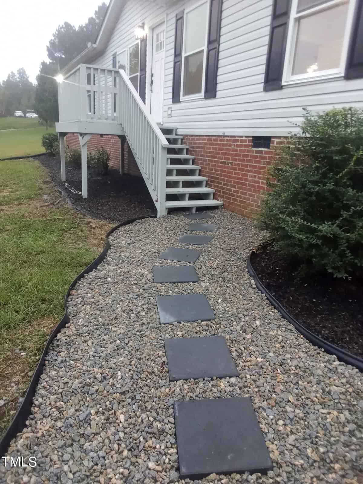 A gravel path with square stepping stones leads to a house with white siding and a small staircase. The path is bordered by black edging and a bush is visible to the right. The house has dark shutters and a red brick foundation.