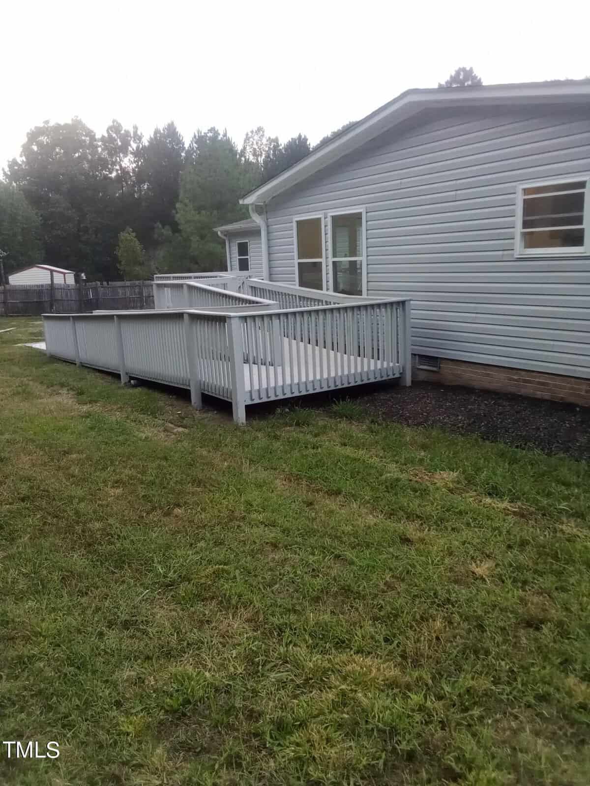A side view of a gray house featuring a wooden ramp with white railings leading to an entrance. The house is surrounded by a grassy yard and bordered by a wooden fence, with trees in the background.