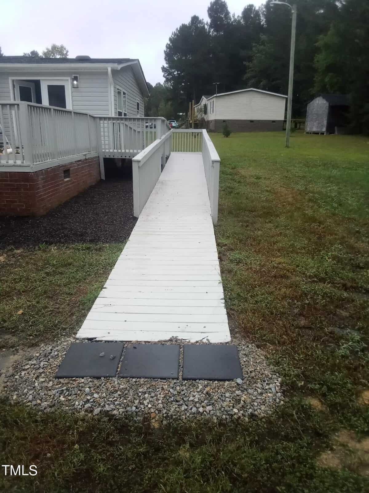 A white wooden ramp leads from a small gravel walkway to a light gray house. The side of the house features a deck with railing. The yard is grassy, bordered by trees in the background, under an overcast sky.