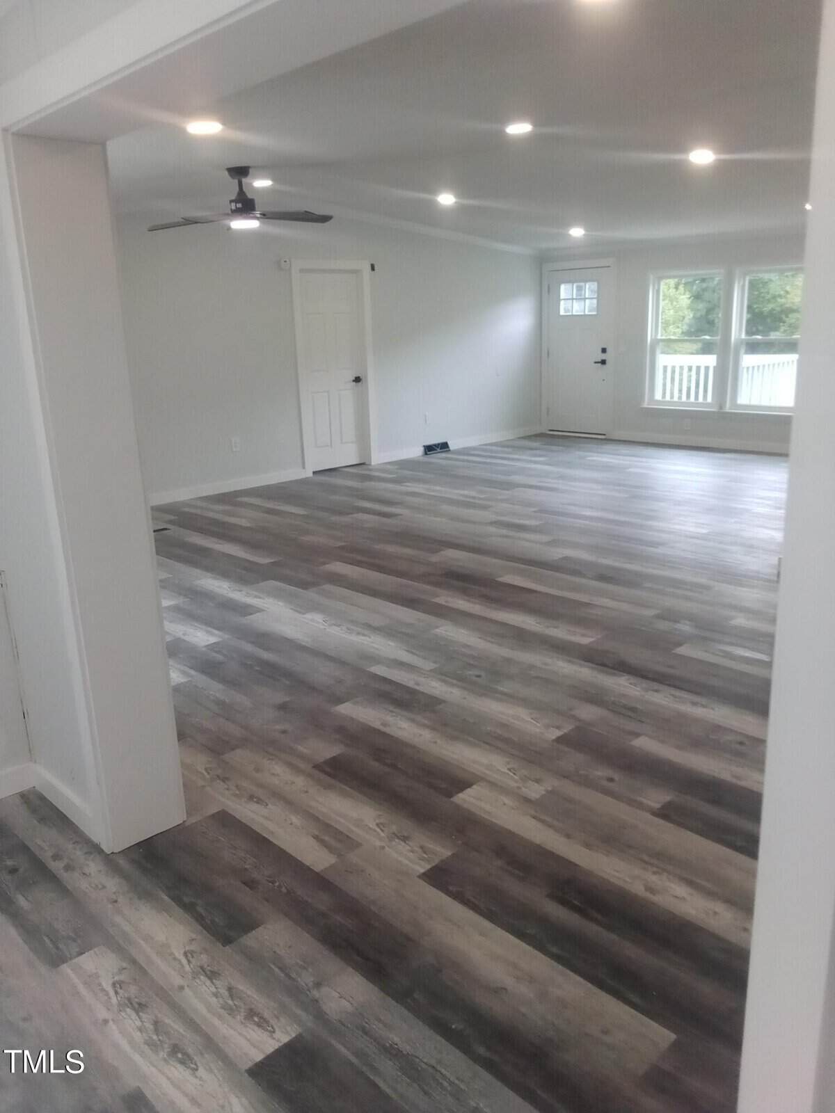 A spacious, empty room with gray wood flooring, white walls, and ceiling lights. There's a ceiling fan, a window, and a door leading outside. The room's open layout and neutral colors create a bright, modern atmosphere.