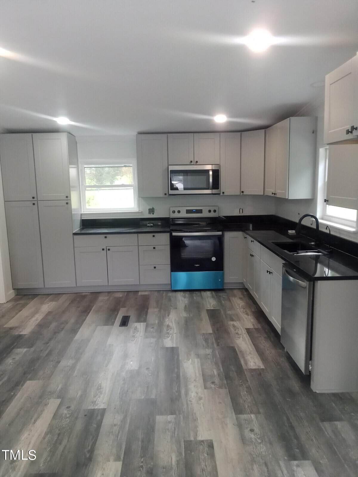 A modern kitchen with white cabinets, stainless steel appliances, and a black countertop. The floor has a wood-like design, and there are two windows providing natural light.
