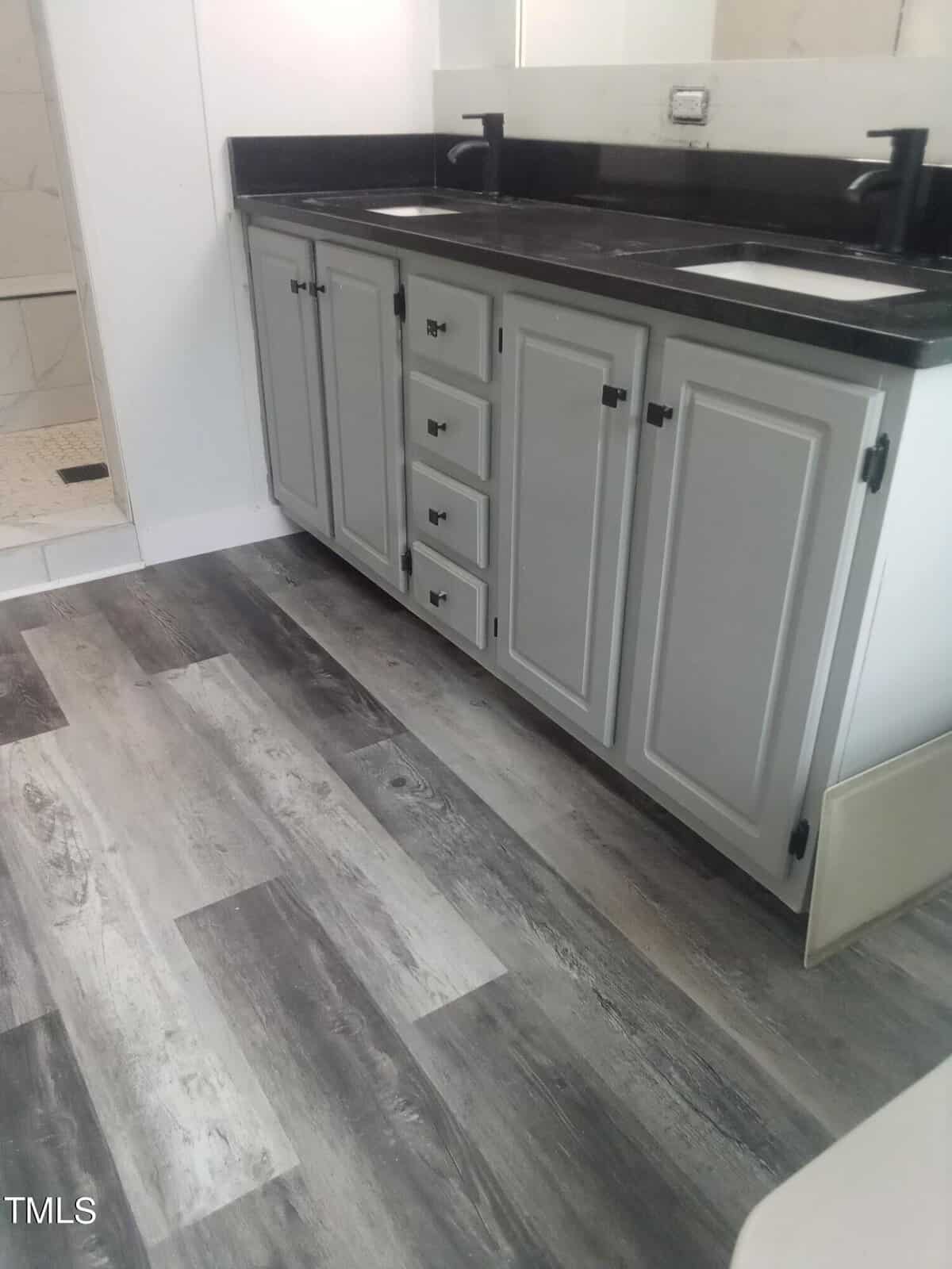 A bathroom with a dual-sink vanity featuring gray cabinets and black countertops. The floor has a wood-like finish in shades of gray and brown. There is a partially visible shower area with light-colored tiles in the background.