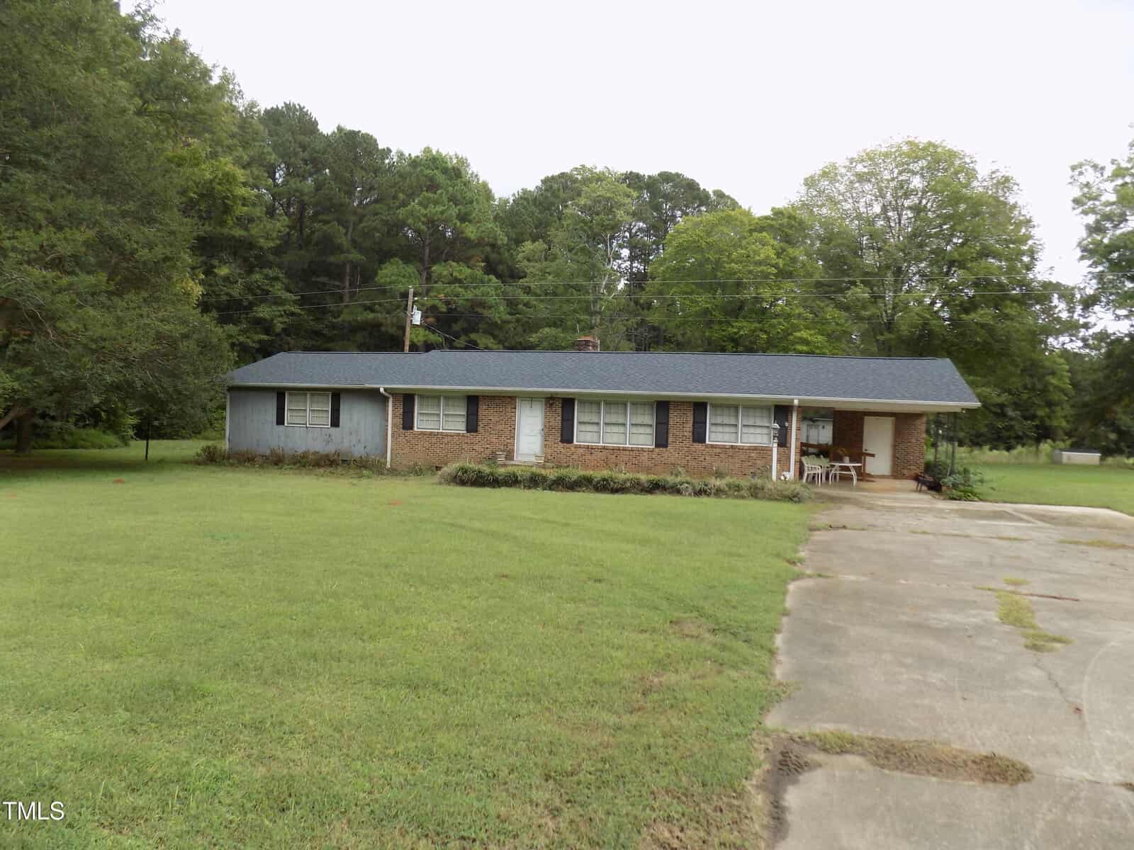 A single-story brick house with a dark shingled roof, a driveway, front porch with chairs, and a large grassy yard. Trees surround the property in the background.