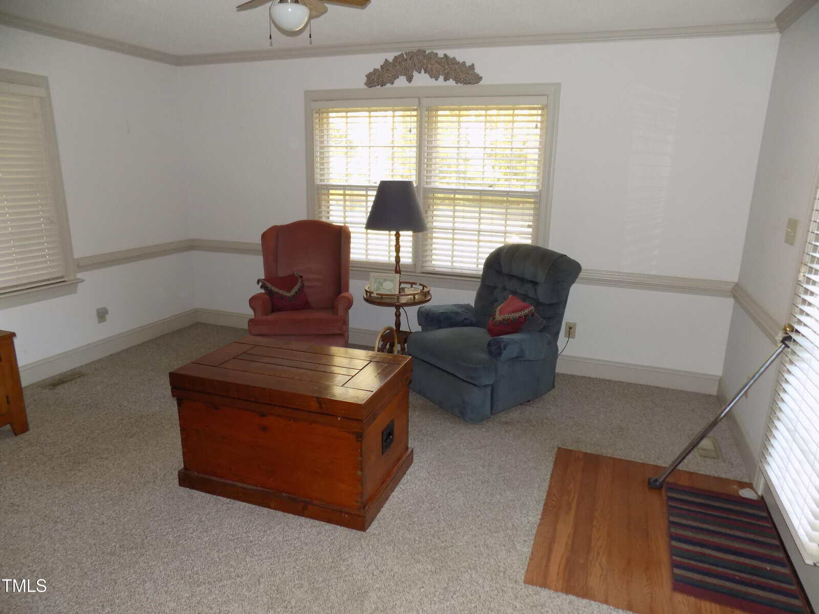 A living room with two armchairs, a wooden chest as a coffee table, a floor lamp, and windows with blinds. A striped doormat and part of a handrail are visible near the wooden floor section.