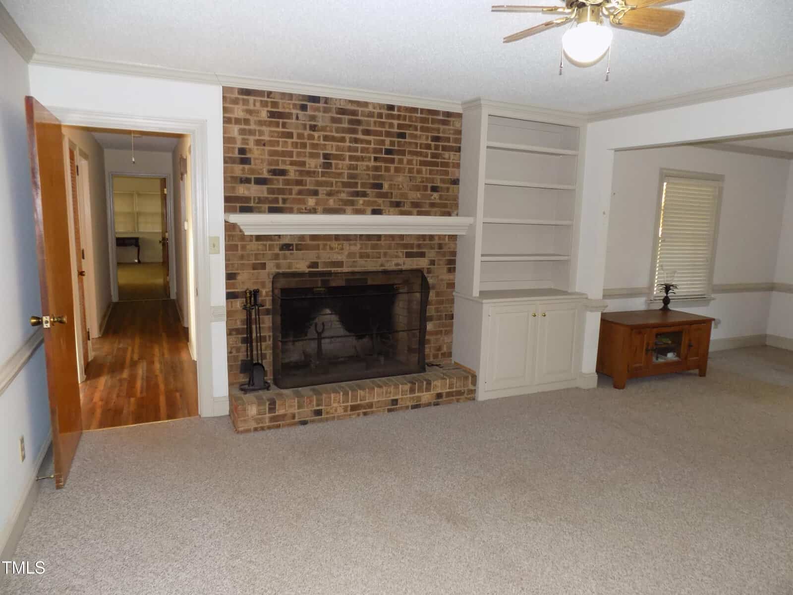 Living room with beige carpet, brick fireplace, white built-in shelves, small ceiling fan, wooden TV stand, and open doorway leading to a hallway with wood flooring.