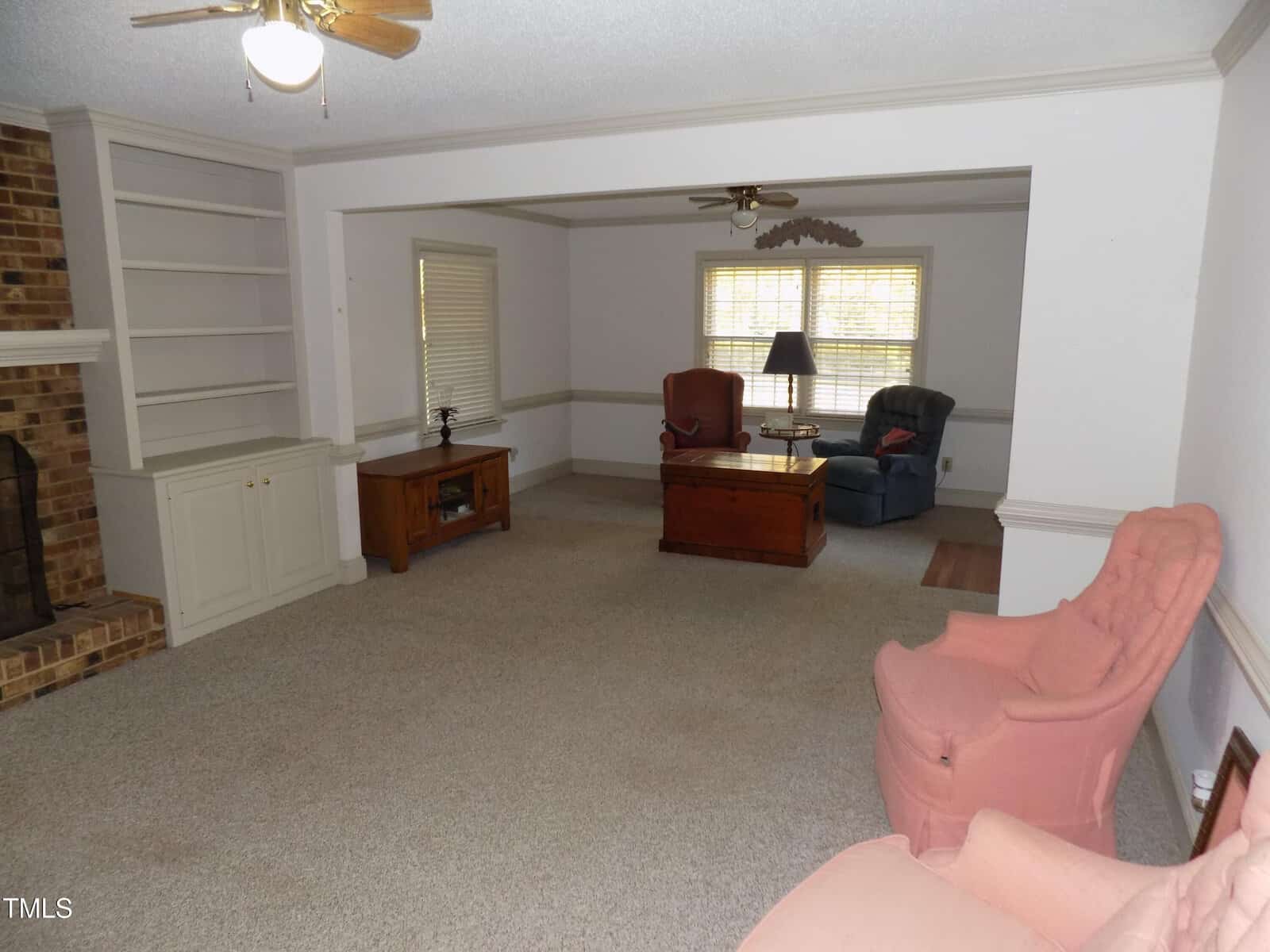 A spacious living room with beige carpet, brick fireplace, built-in shelves, a TV stand, two armchairs, a lamp on a chest, and large windows letting in natural light.