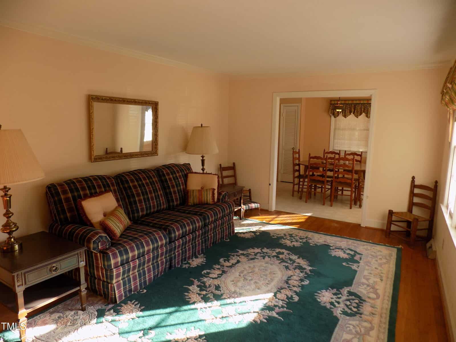 A living room with a plaid sofa, end tables with lamps, a large oval rug, and a wall mirror. Wooden chairs line the walls. An open doorway leads to a dining area with a table and chairs. Sunlight streams in from a window.