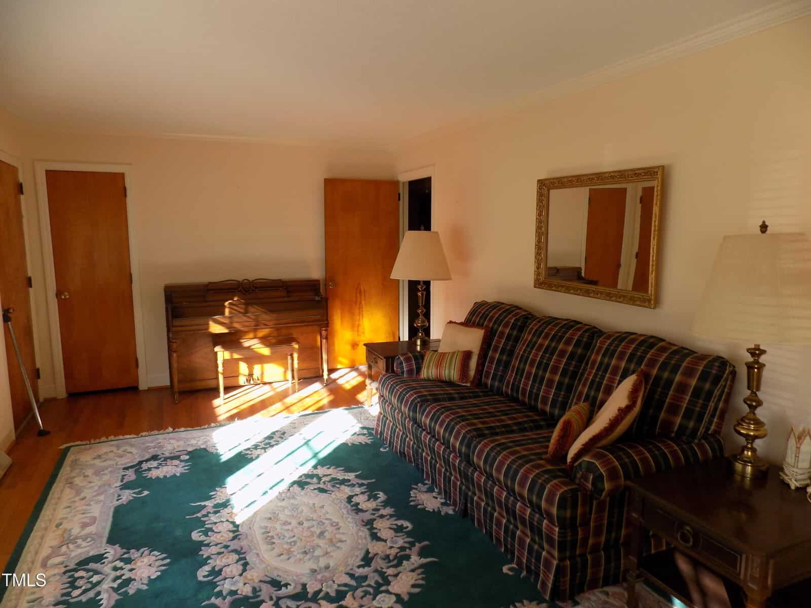 A living room with a plaid sofa, two lamps, a large decorative rug, a mirror on the wall, a small side table, and sunlight streaming in from a window. There are two wooden doors and a closed upright piano.