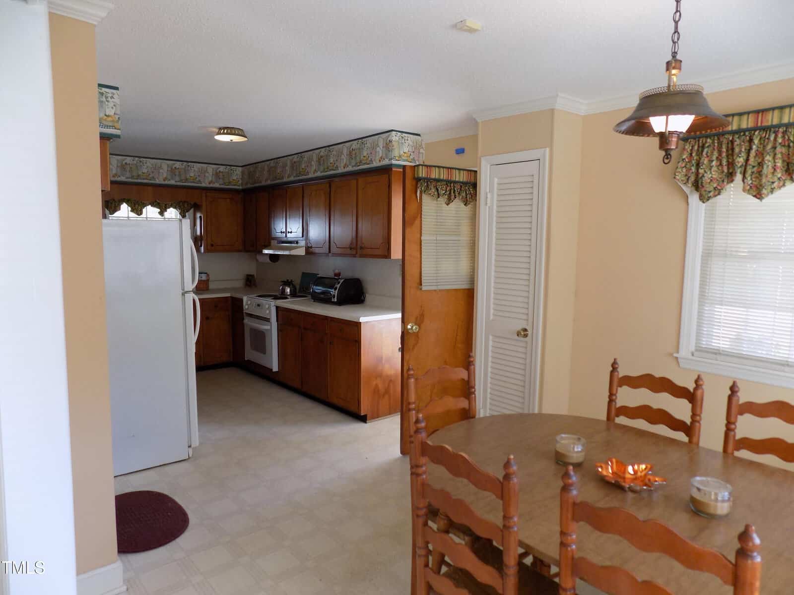 A kitchen and dining area with wood cabinets, white appliances, a round wooden table with four chairs, beige walls, and floral window valances. The kitchen has a refrigerator, stove, and countertop appliances.
