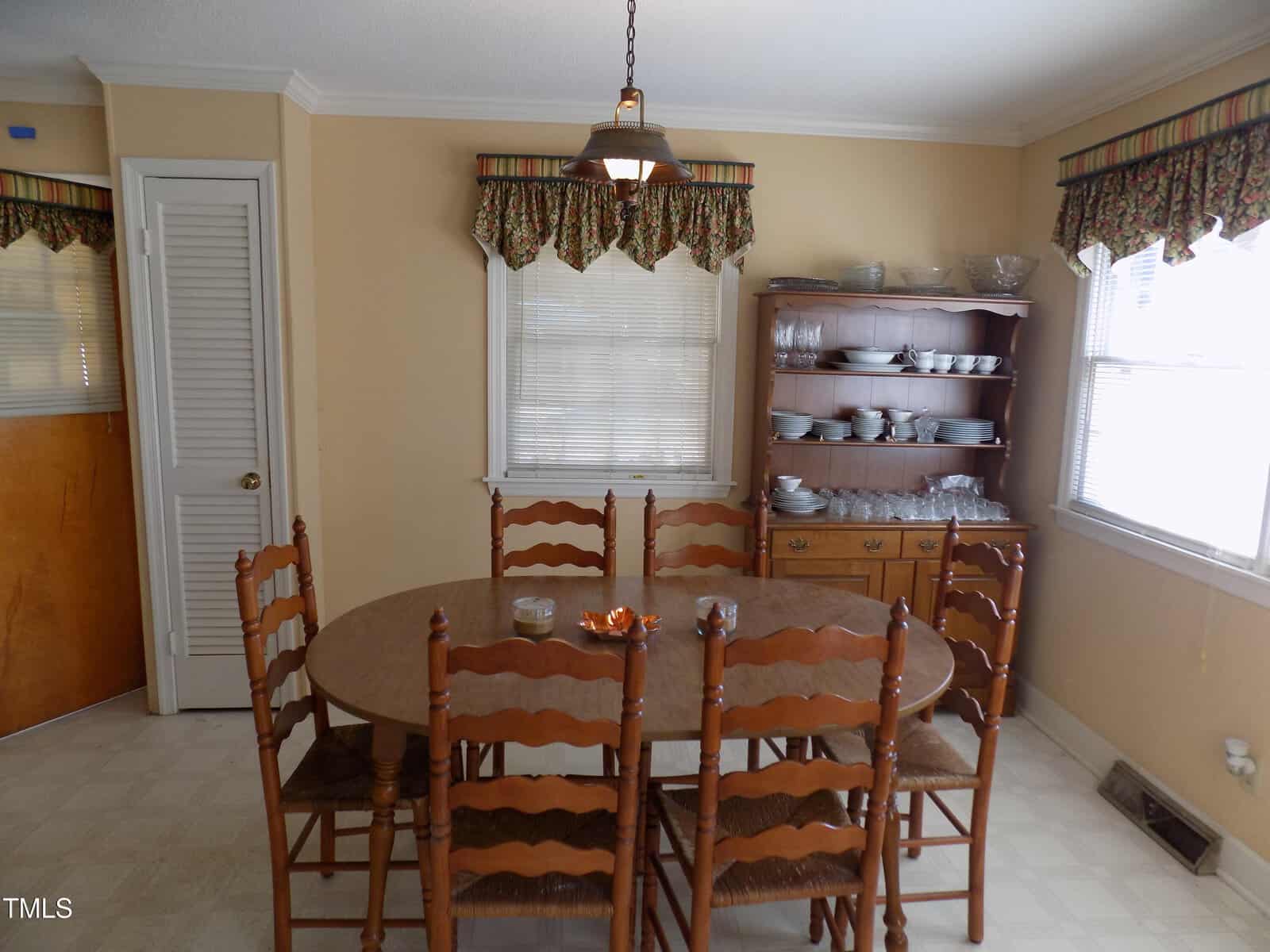 A dining room with a wooden oval table and six matching chairs, a wooden hutch with dishes and glassware, beige walls, two windows with patterned valances, and a ceiling light fixture above the table.