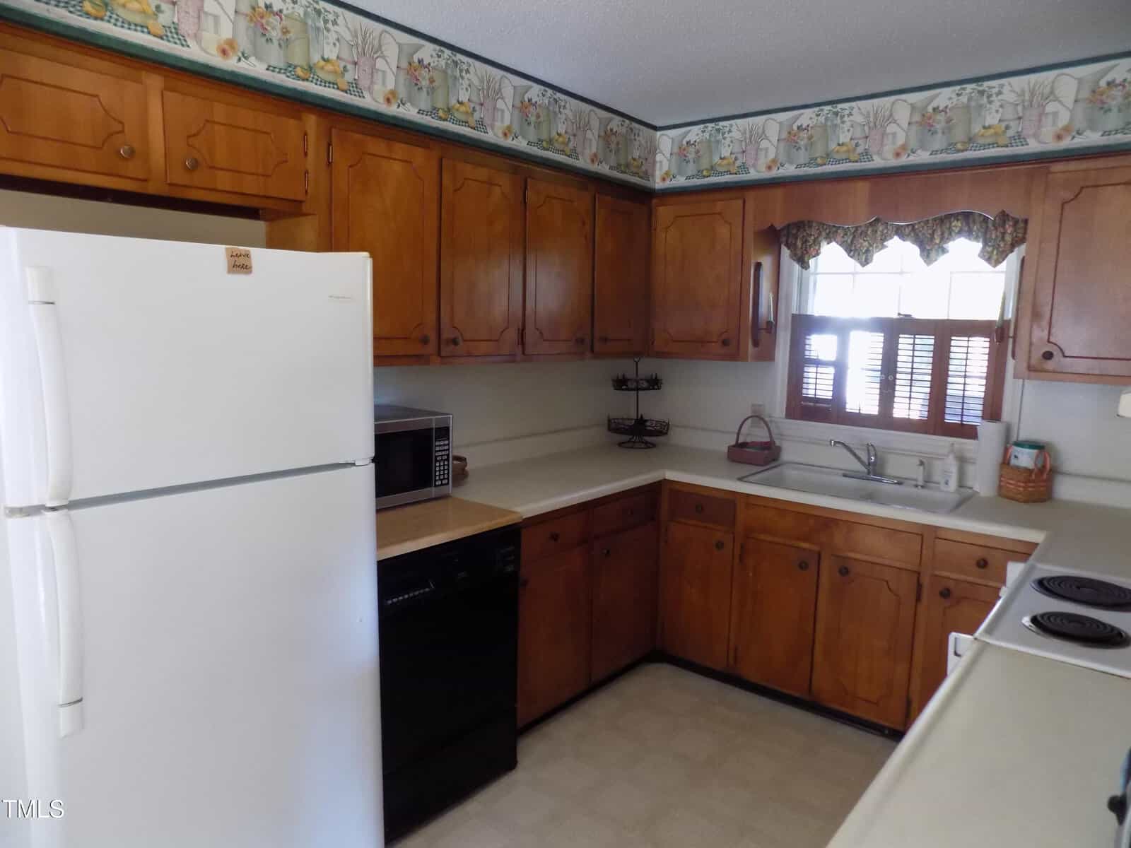 A kitchen with wood cabinets, a white refrigerator, a black dishwasher, a microwave, a sink under a window with a patterned valance, light countertops, and wallpaper featuring a floral and basket design along the top.
