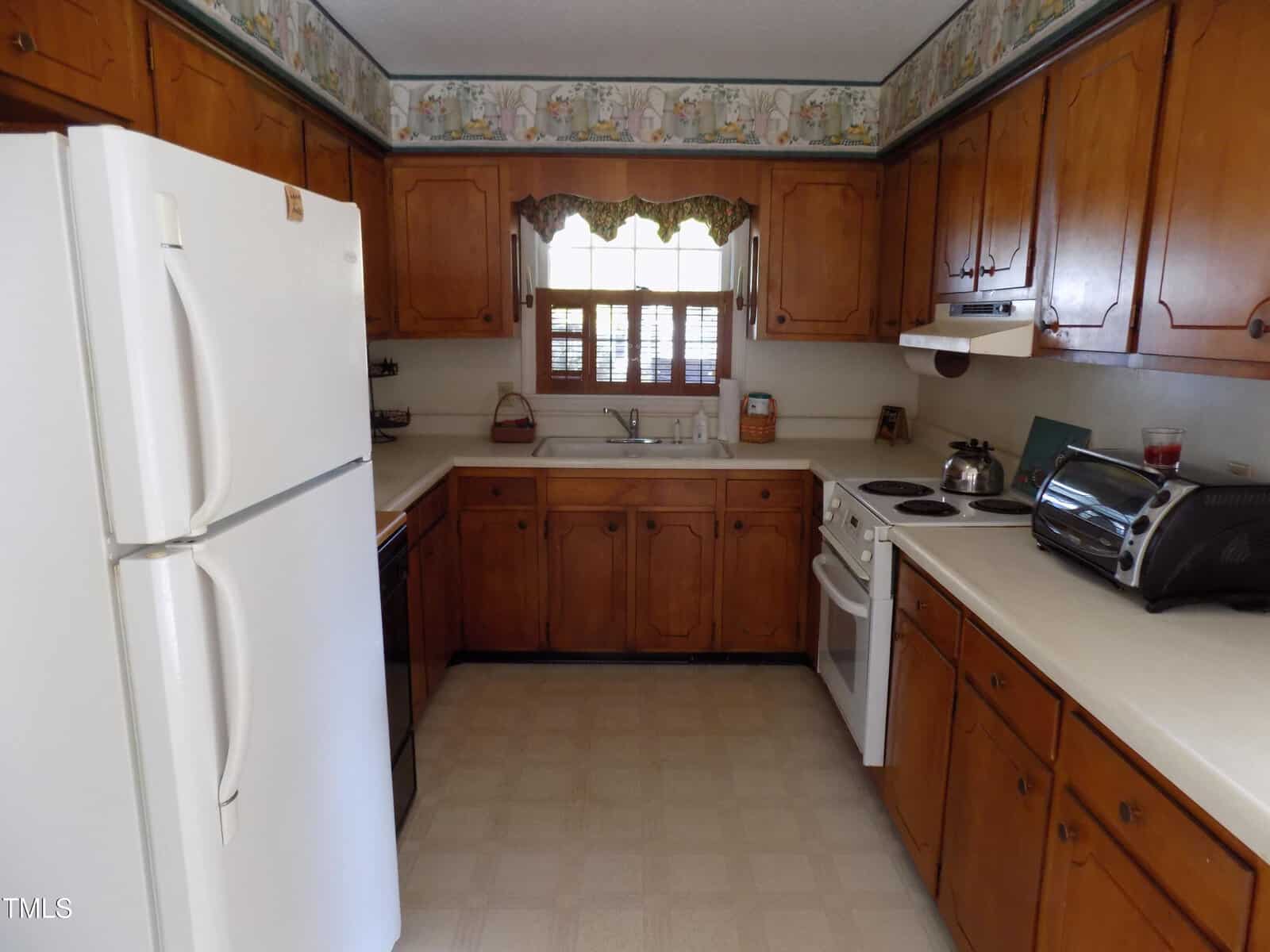 A traditional kitchen with wood cabinets, white appliances including a refrigerator and stove, a double sink under a window with a scalloped valance, and patterned wallpaper border near the ceiling.
