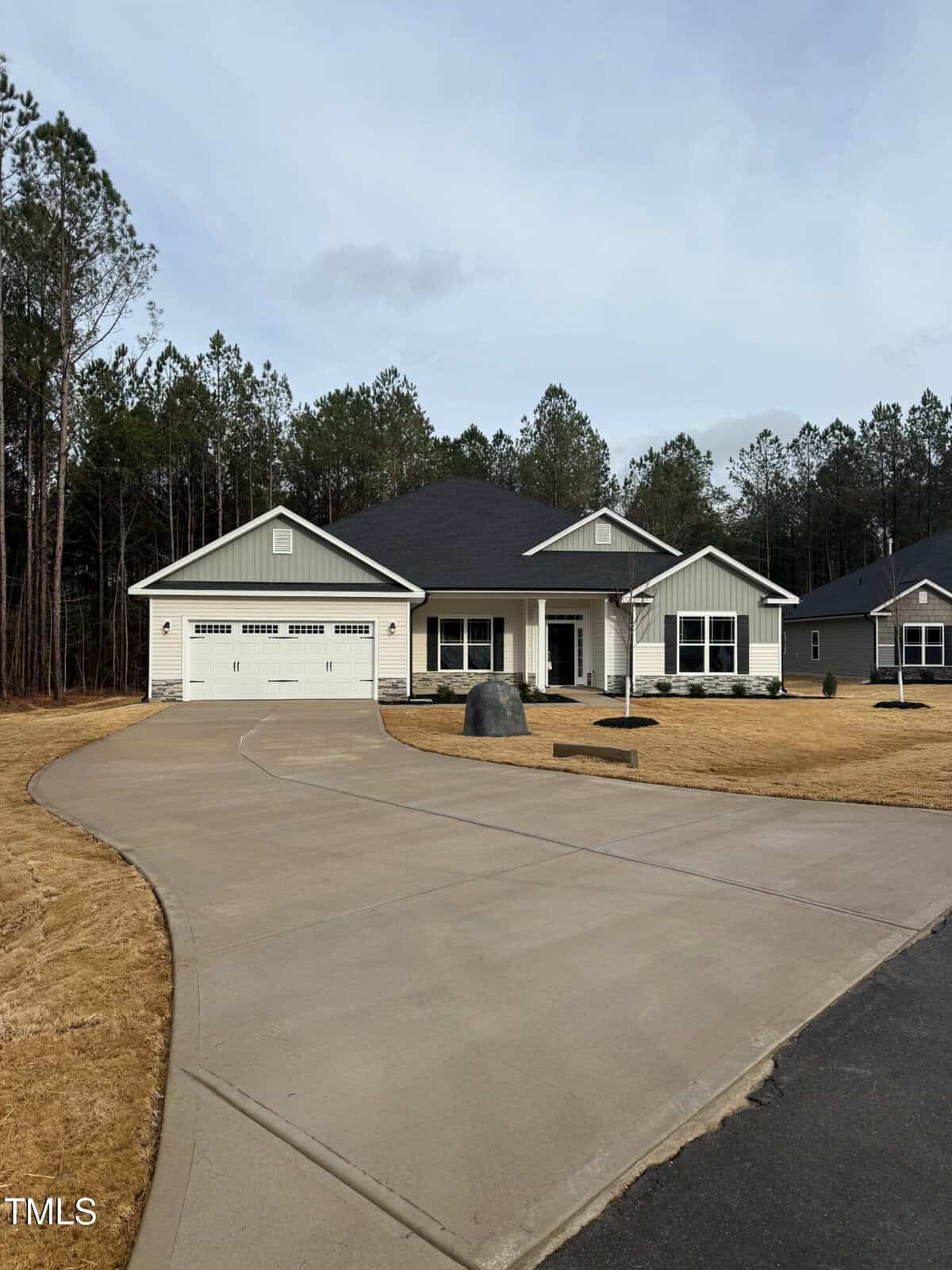 Single-story house with a dark roof, light siding, and attached two-car garage. The house has a wide driveway, front porch, minimal landscaping, and is bordered by trees in the background.