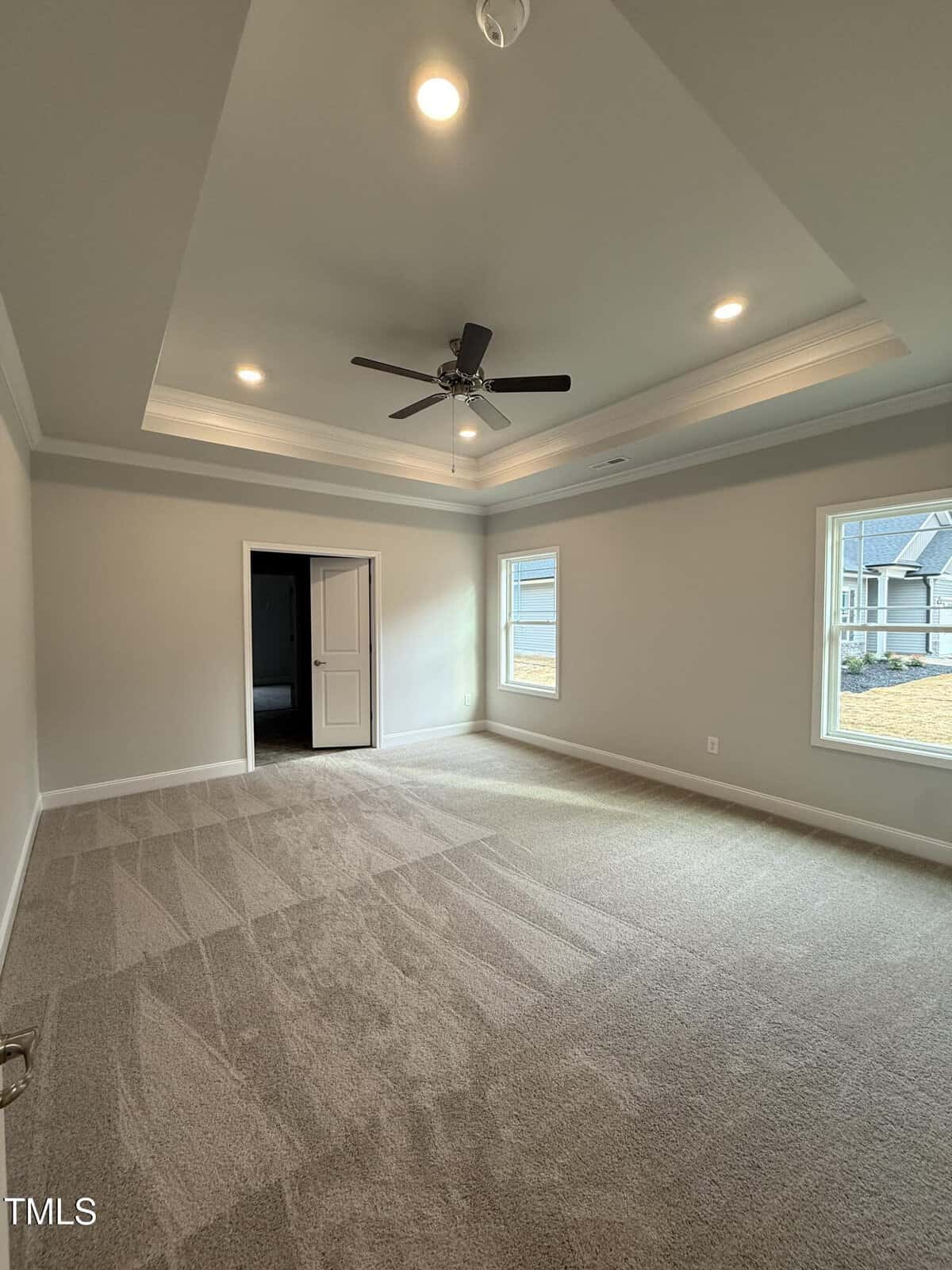 A spacious, empty room with beige carpet, light gray walls, a tray ceiling with recessed lighting, a ceiling fan, two windows, and an open door leading to another room.