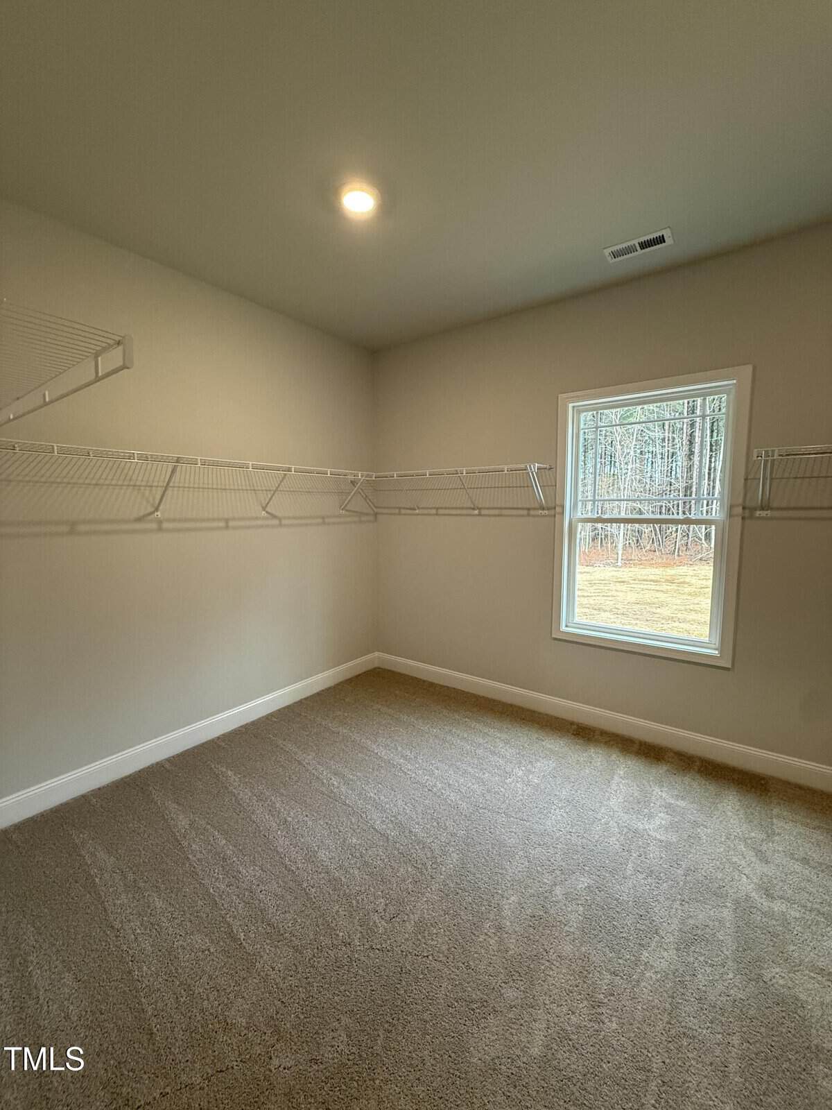 A small empty room with beige carpet, light-colored walls, a single window, and white wire shelving installed along two walls. A ceiling light is turned on, and the view outside shows trees and grass.