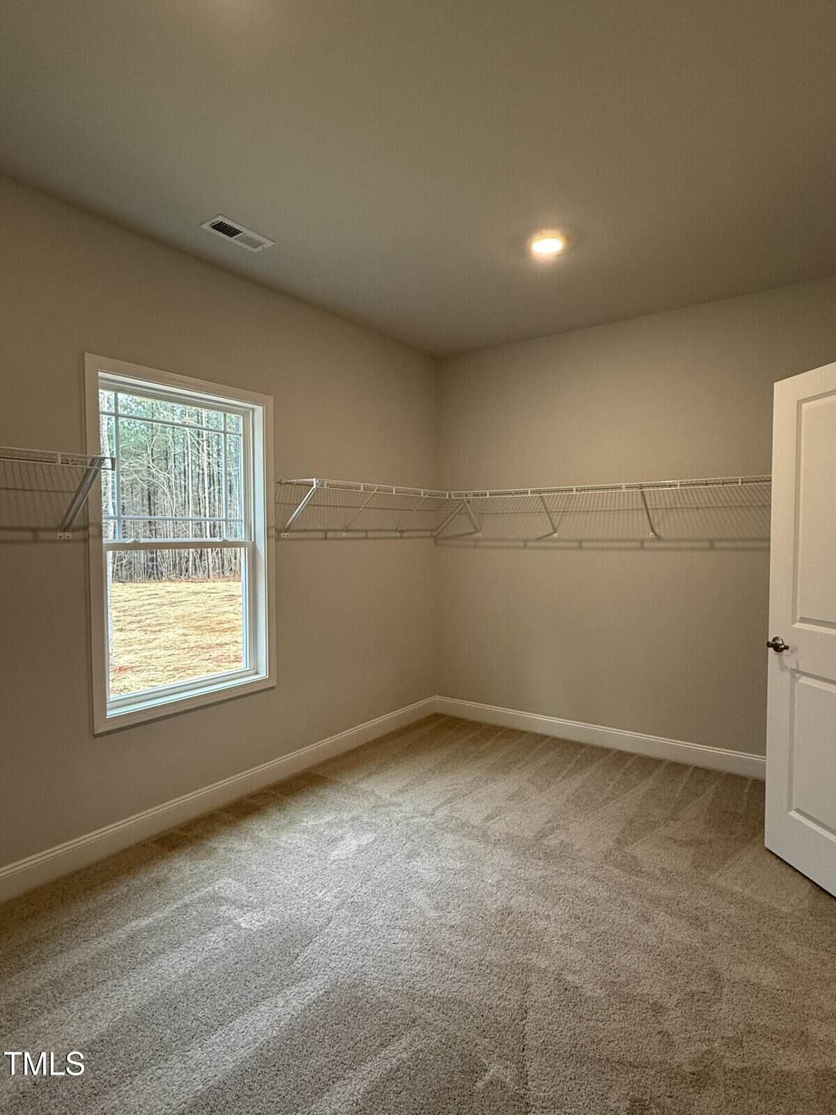 A small, empty walk-in closet with beige carpet, light tan walls, wire shelving along two walls, a single window, and an open white door. A recessed ceiling light illuminates the room.