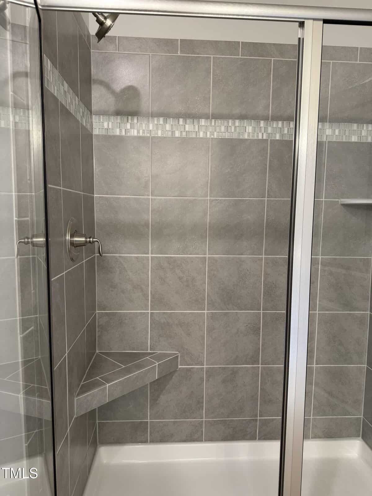 Modern shower with gray tile walls, a glass door, a corner bench, and a decorative horizontal strip of small square tiles near the top. The showerhead and control handle are silver.
