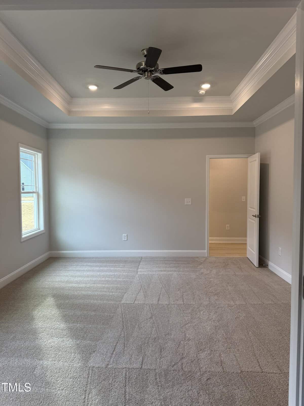 Empty room with beige carpet, light gray walls, a tray ceiling with recessed lights and a ceiling fan, one window on the left, and an open door leading to another area with different flooring.