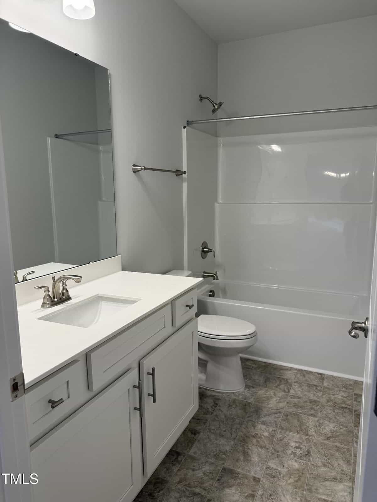 Modern bathroom with white cabinets, a large mirror, a white countertop with a sink, a toilet, and a bathtub-shower combo with chrome fixtures. The floor has gray patterned tiles.