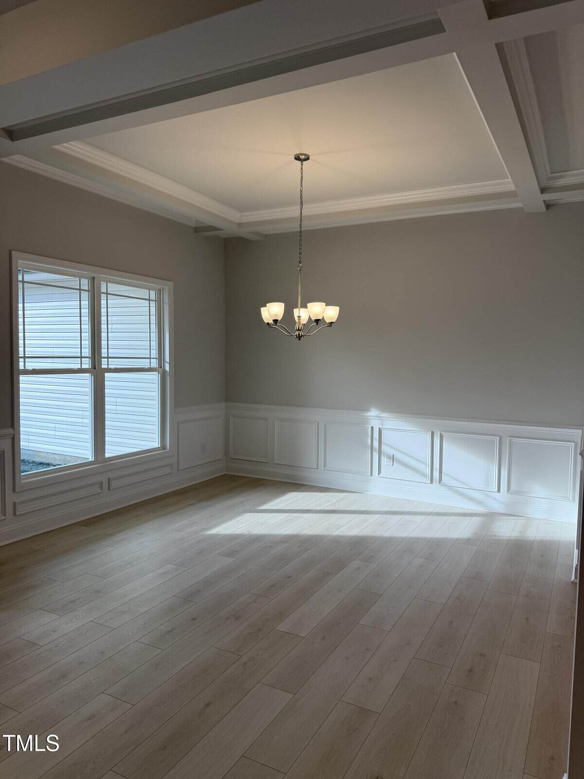 A bright, empty dining room with light wood floors, gray walls, white wainscoting, large windows, a coffered ceiling, and a chandelier hanging in the center. Sunlight casts shadows on the floor and wall.