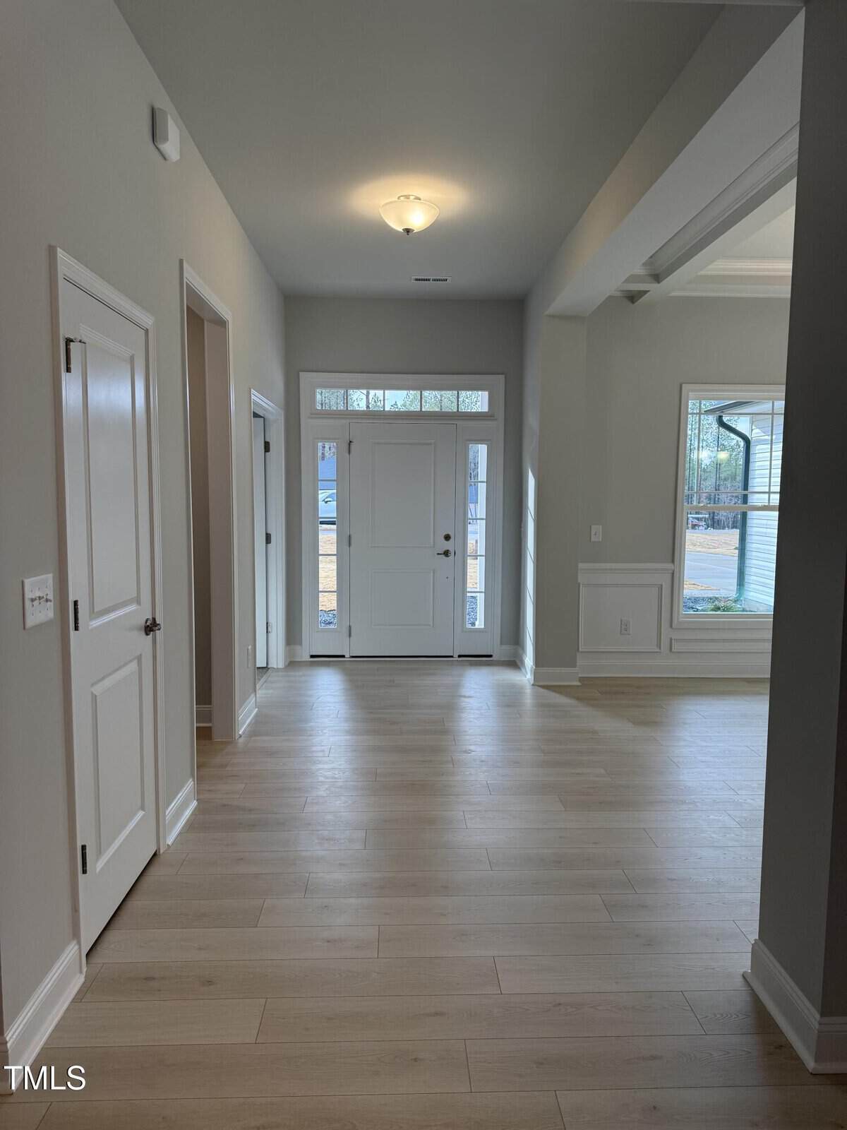 A hallway with light wood flooring, neutral walls, and white trim. There are two closed doors on the left and a front door with glass panels and sidelights at the end. Sunlight comes in through the windows.