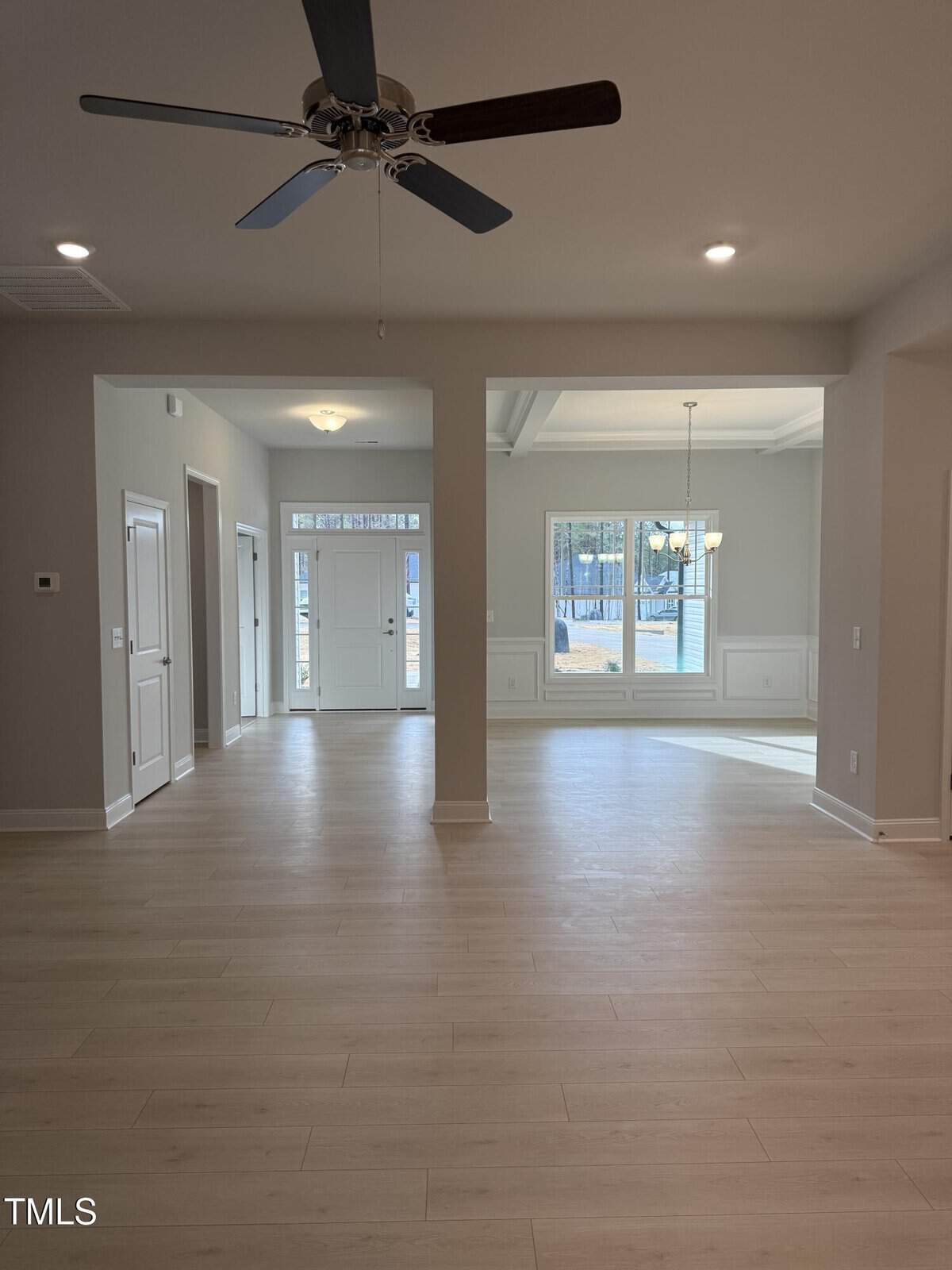 A spacious, empty room with light wood flooring, a ceiling fan, recessed lighting, and a view of a white front door and large window with a hanging light fixture over a dining area.