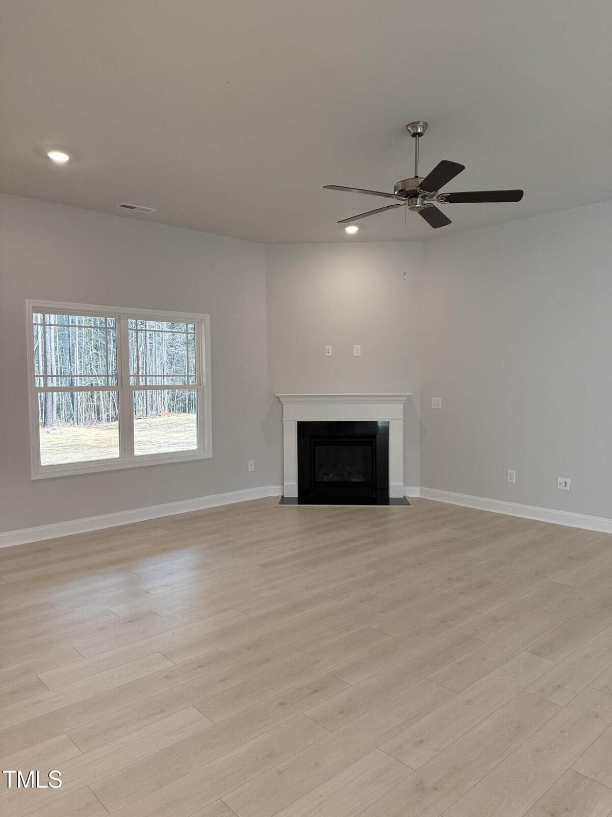 Empty living room with light wood flooring, gray walls, a white-framed window, a corner fireplace with a white mantel, and a ceiling fan with lights. Natural light enters through the window.