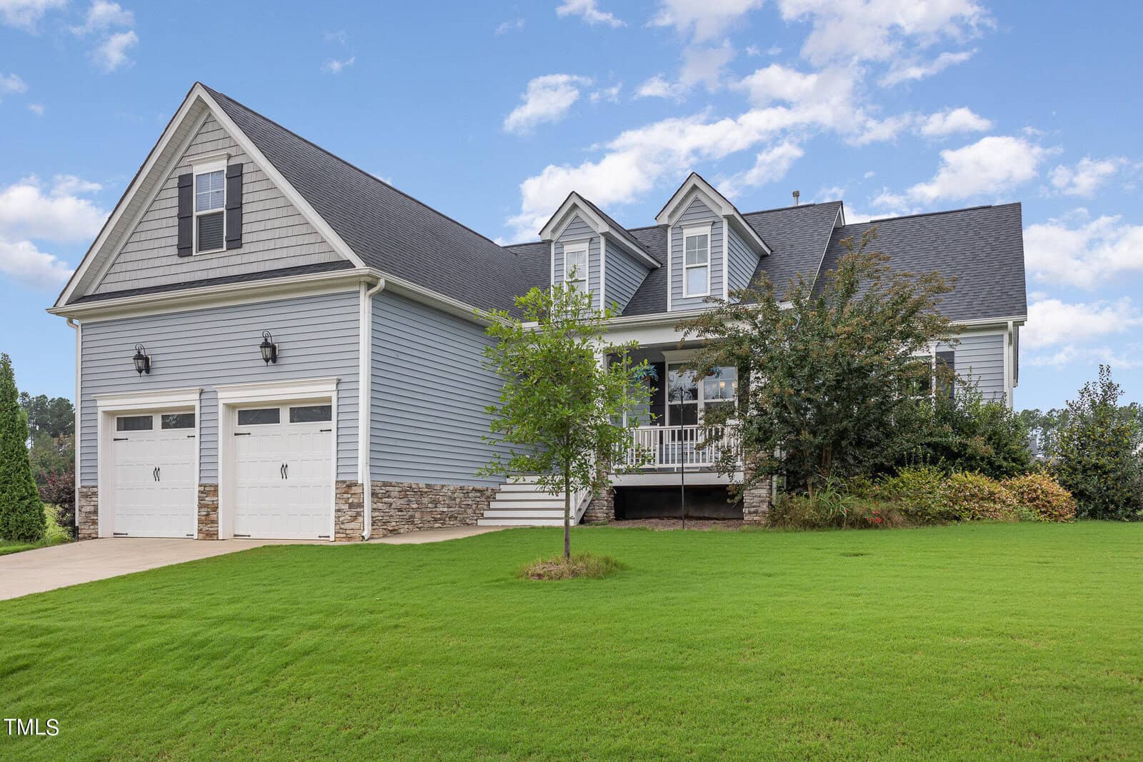 A two-story suburban house with gray siding, white trim, a double garage, front porch, dormer windows, and a neatly manicured green lawn under a partly cloudy sky.