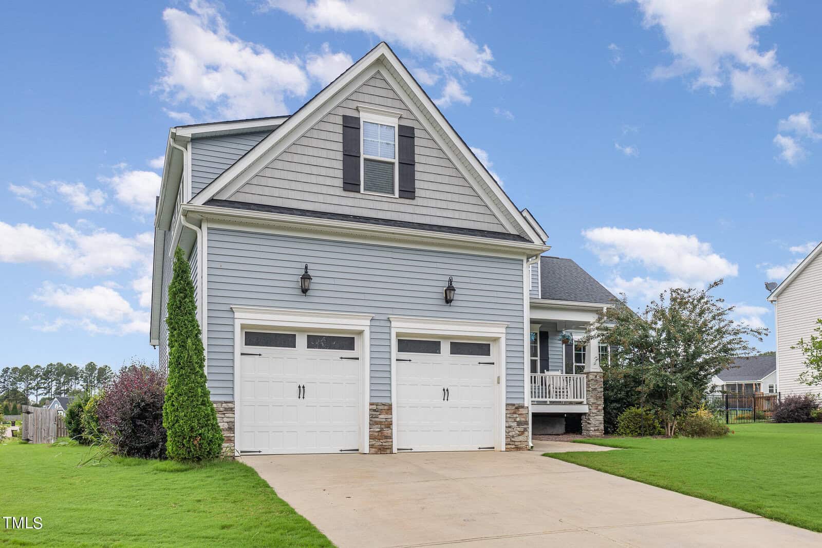 A two-story suburban house with light gray siding, white trim, and two garage doors. The driveway is concrete, and the front yard has green grass and shrubs. The sky is blue with scattered clouds.