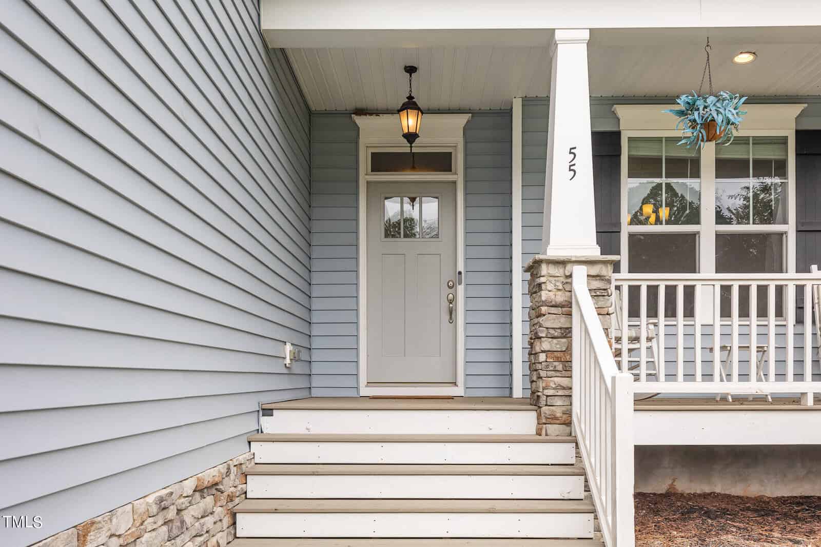 Front porch of a house with gray siding, a white door with glass panes, stone column, white railings, steps leading up, a hanging lantern, and a blue decorative plant to the right. House number 55 is visible on the post.
