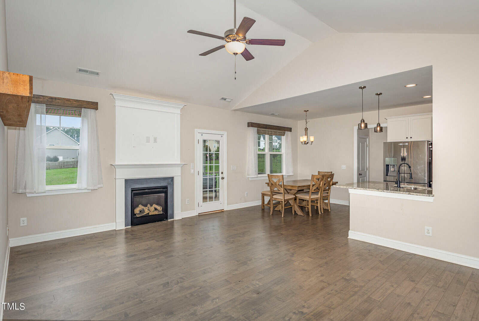 Open living space with wood floors, a ceiling fan, fireplace, and large windows. The area includes a dining table near the kitchen, which features white cabinets, stainless steel appliances, and pendant lighting.