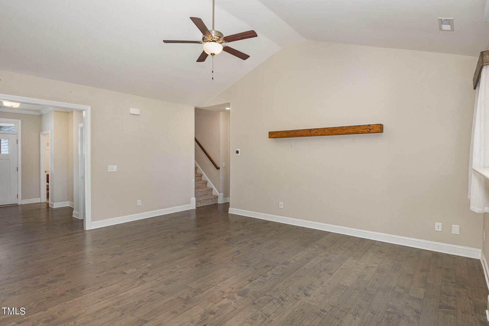 Spacious living room with hardwood floors, beige walls, a ceiling fan with light, and a wooden shelf on the wall. Stairs and a hallway leading to other rooms are visible in the background.