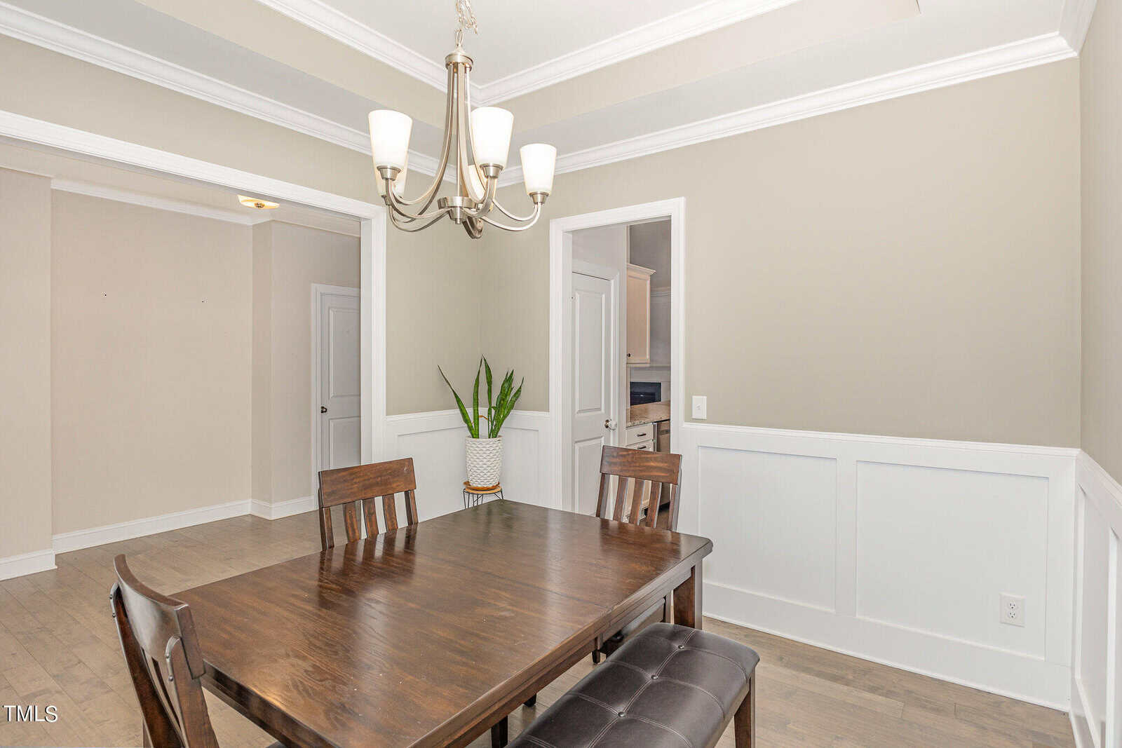 A dining room with a dark wooden table, three chairs, and a bench. A potted plant sits on the table. The walls are light with white wainscoting, and a chandelier hangs from the ceiling. Doorways lead to adjoining rooms.