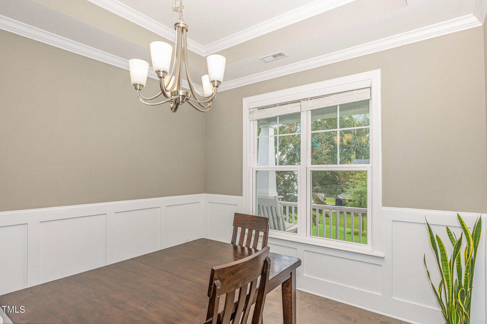 A dining room with a wooden table and chairs, beige walls with white wainscoting, a modern chandelier, a large window with blinds, and a green potted plant in the corner.
