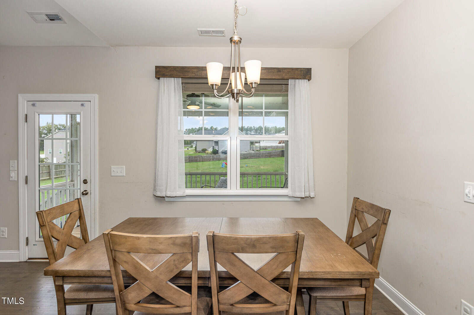 A dining area with a wooden table and four matching chairs, set by a window with white curtains. A light fixture hangs above the table, and a backyard with a fence is visible through the window.