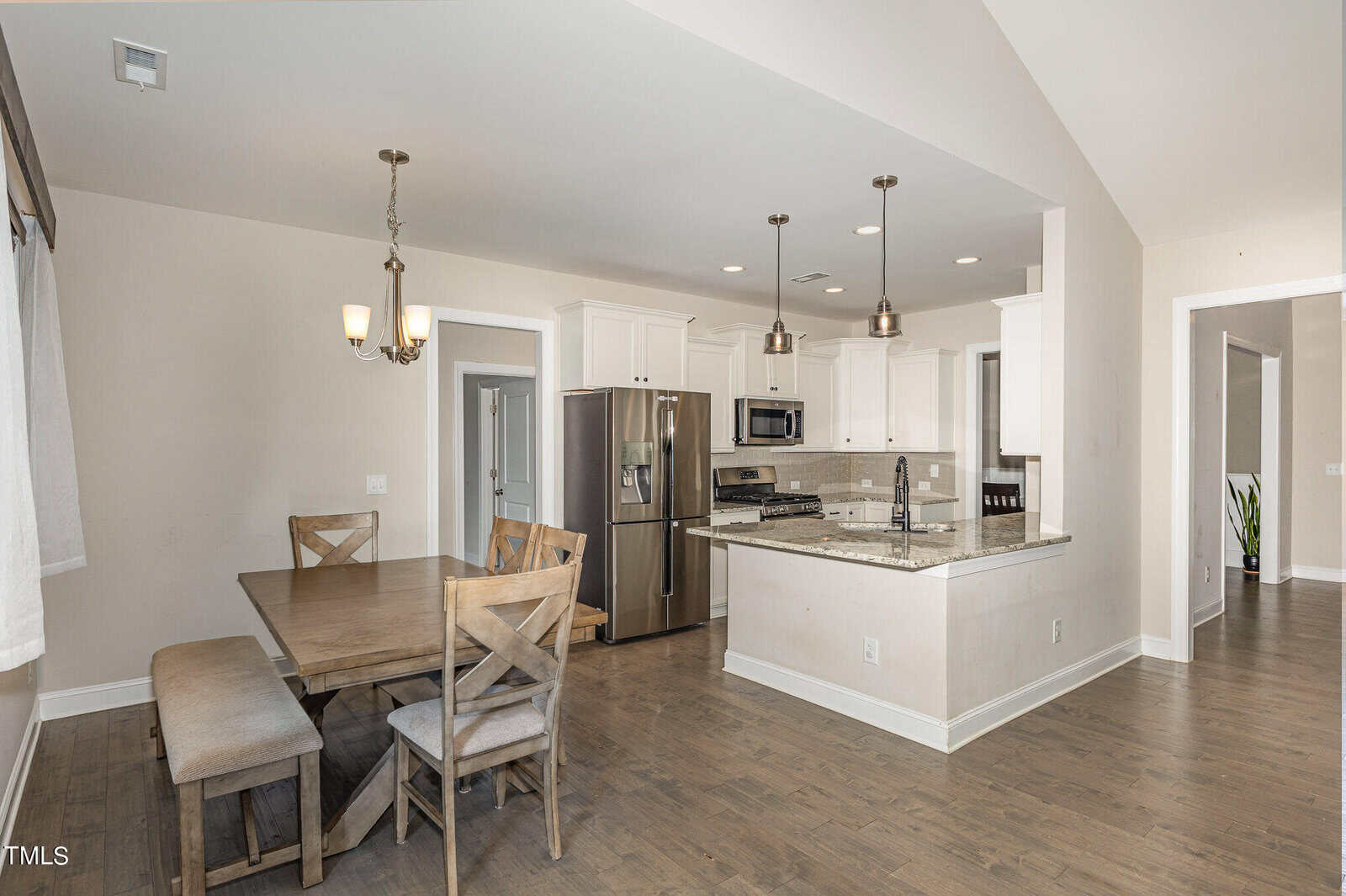 A modern kitchen and dining area with wooden floors, stainless steel appliances, white cabinets, granite countertops, pendant lighting over the island, and a wooden dining table with chairs and a bench.