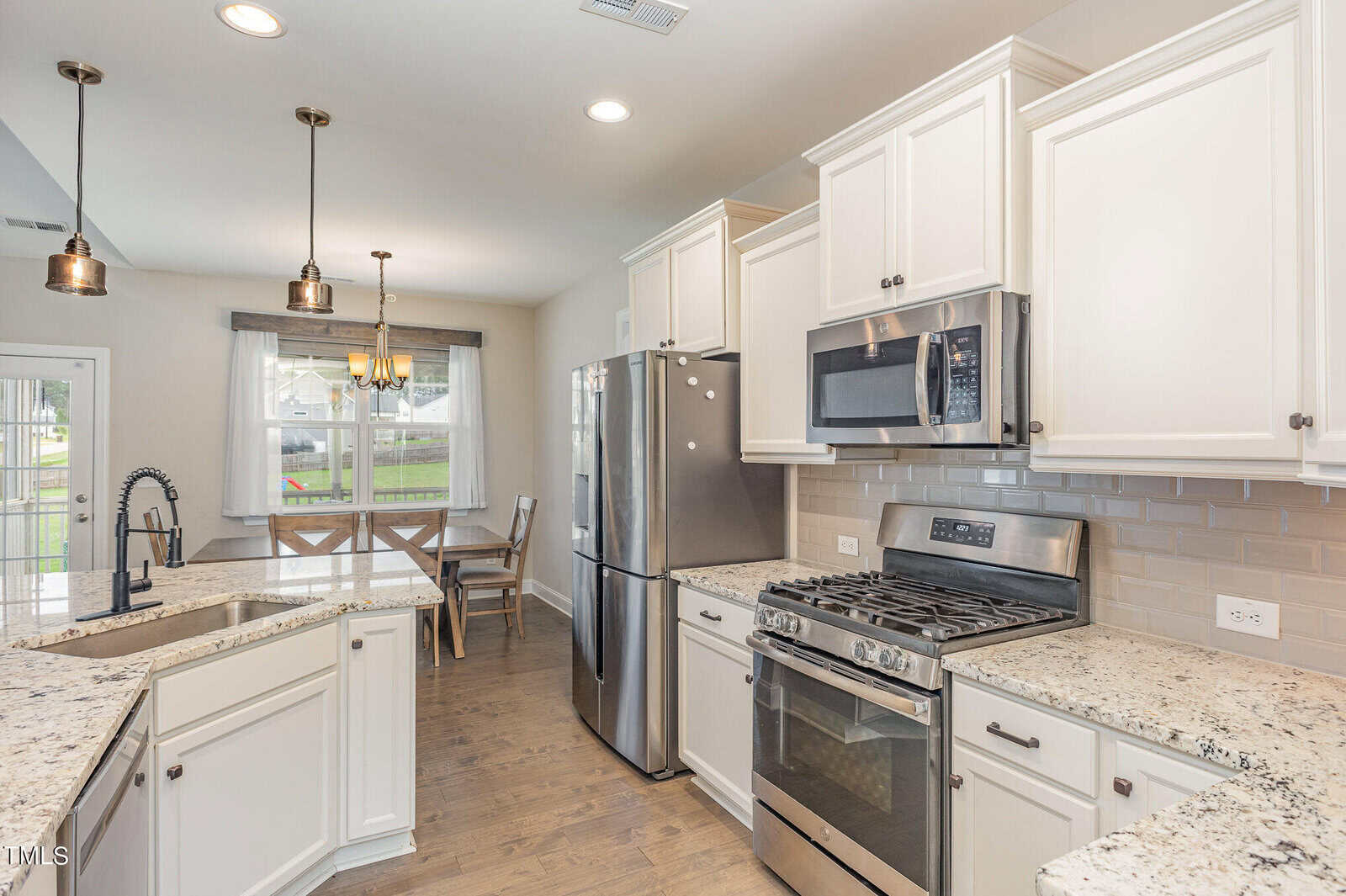 Modern kitchen with white cabinets, stainless steel appliances, granite countertops, and a dining area by a window. Pendant lights hang above the island, and a subway tile backsplash lines the wall behind the stove and microwave.