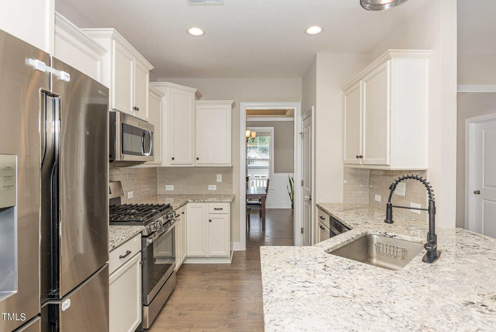 Modern kitchen with stainless steel appliances, white cabinets, granite countertops, and a tile backsplash. A gooseneck faucet is installed on the island. A dining area is visible through a doorway in the background.