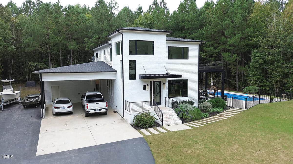 Modern two-story white house with large windows, a carport sheltering two vehicles, and a backyard pool, surrounded by trees and a spacious yard with landscaped greenery.