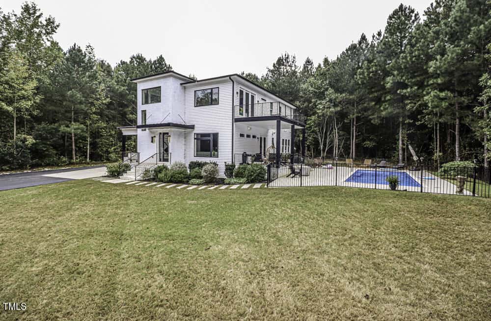 Modern two-story white house with large windows, a balcony, and a covered porch, surrounded by trees. There is a fenced-in swimming pool and landscaped yard with a grassy lawn in the foreground.