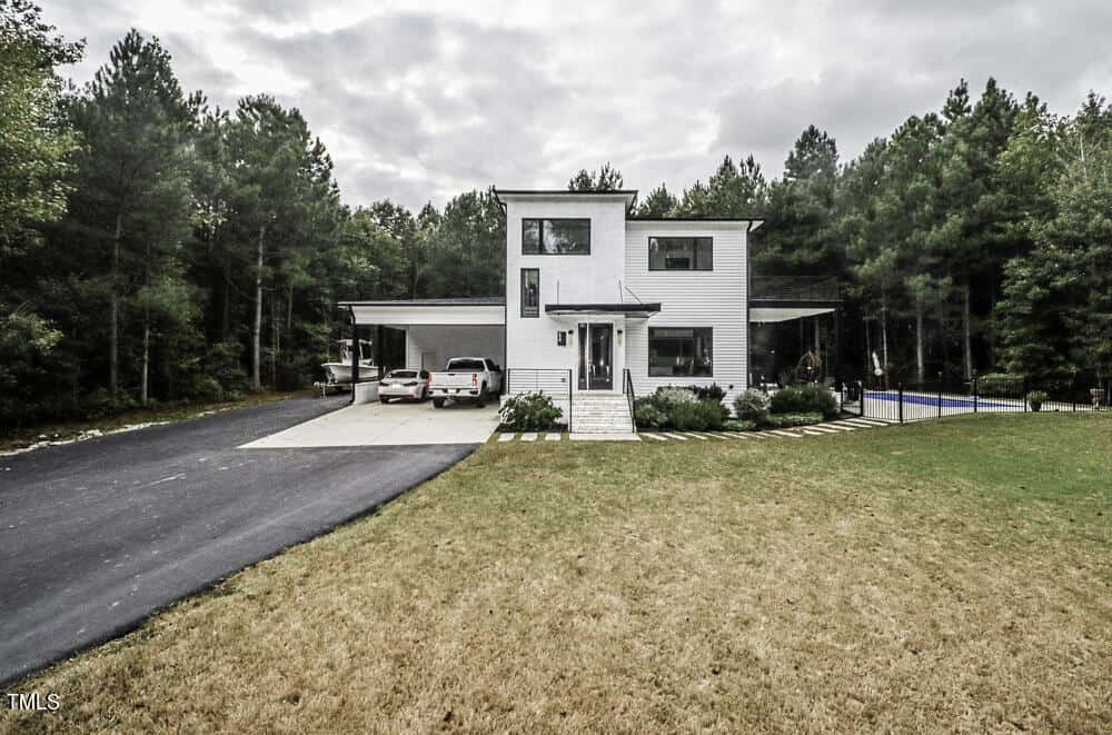 A modern two-story white house with large windows, a carport with two vehicles, and a black driveway, surrounded by trees and a grassy yard under a cloudy sky. A blue pool is visible on the right side.