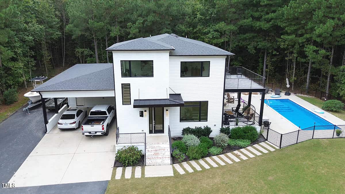 A modern two-story white house with a carport, two vehicles parked, a fenced backyard with a rectangular swimming pool, patio furniture, and surrounded by trees.