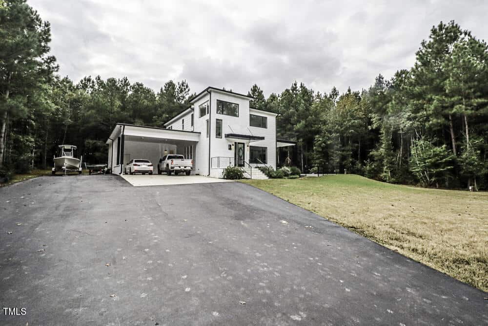 Modern two-story white house with large windows, a covered carport with two parked vehicles, a driveway, boat, and surrounded by trees and a grassy yard under a cloudy sky.