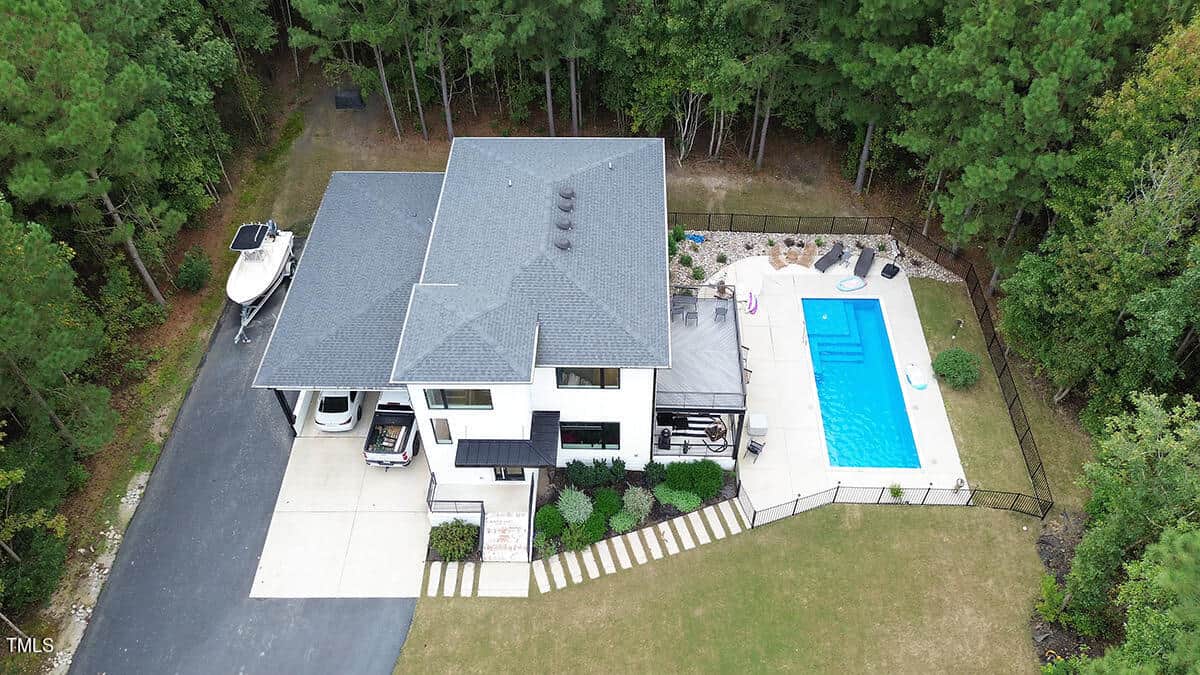 Aerial view of a modern two-story house with a driveway, parked boat, a fenced rectangular swimming pool, patio area, and surrounded by dense green trees.