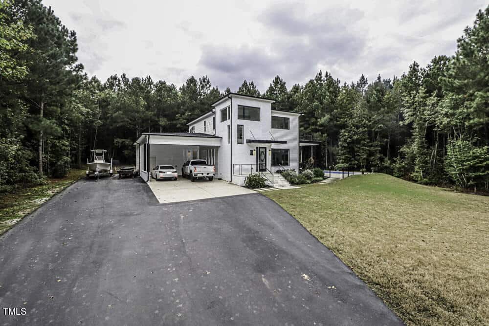 A modern two-story house with white exterior, large windows, and a driveway leading to a carport. Two cars and a boat are parked beside the home, surrounded by trees and a grassy yard.