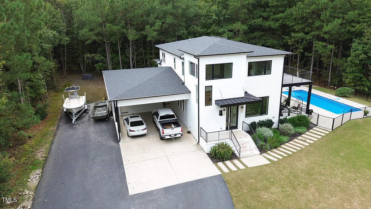 A modern two-story white house with a black roof, two cars parked under a carport, a boat on a trailer, a swimming pool in the backyard, and trees surrounding the property.